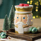 Beleuchtete Nutcracker-Duftlampe mit roten Wachswürfeln zwischen kleinen Tannen und Christbaumkugeln auf einem Holzbrett.
