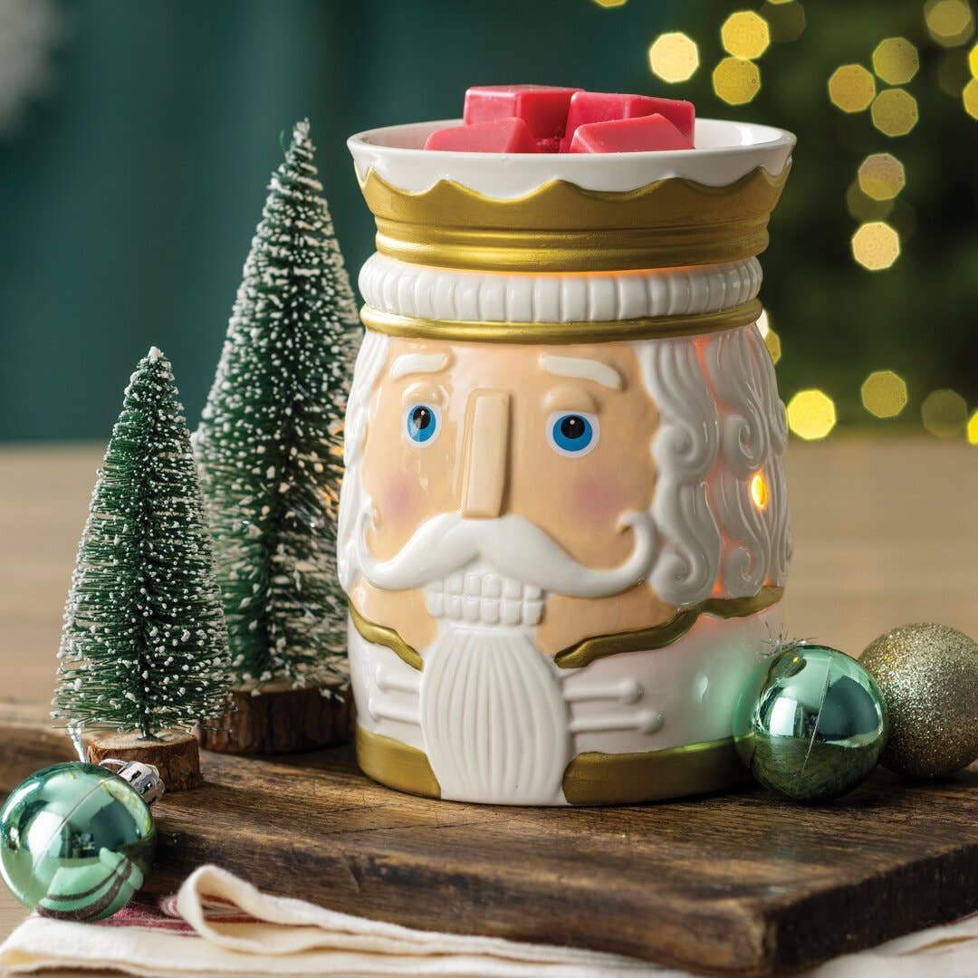 Beleuchtete Nutcracker-Duftlampe mit roten Wachswürfeln zwischen kleinen Tannen und Christbaumkugeln auf einem Holzbrett.