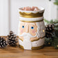 Nutcracker-Duftlampe in Weiß und Gold mit rosafarbenen Wachswürfeln, umgeben von Tannenzapfen und grünen Zweigen.