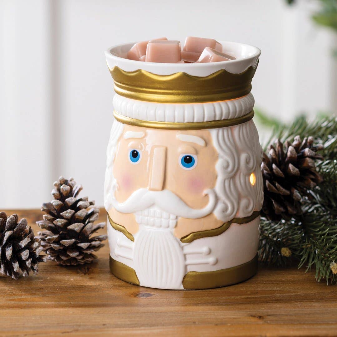 Nutcracker-Duftlampe in Weiß und Gold mit rosafarbenen Wachswürfeln, umgeben von Tannenzapfen und grünen Zweigen.